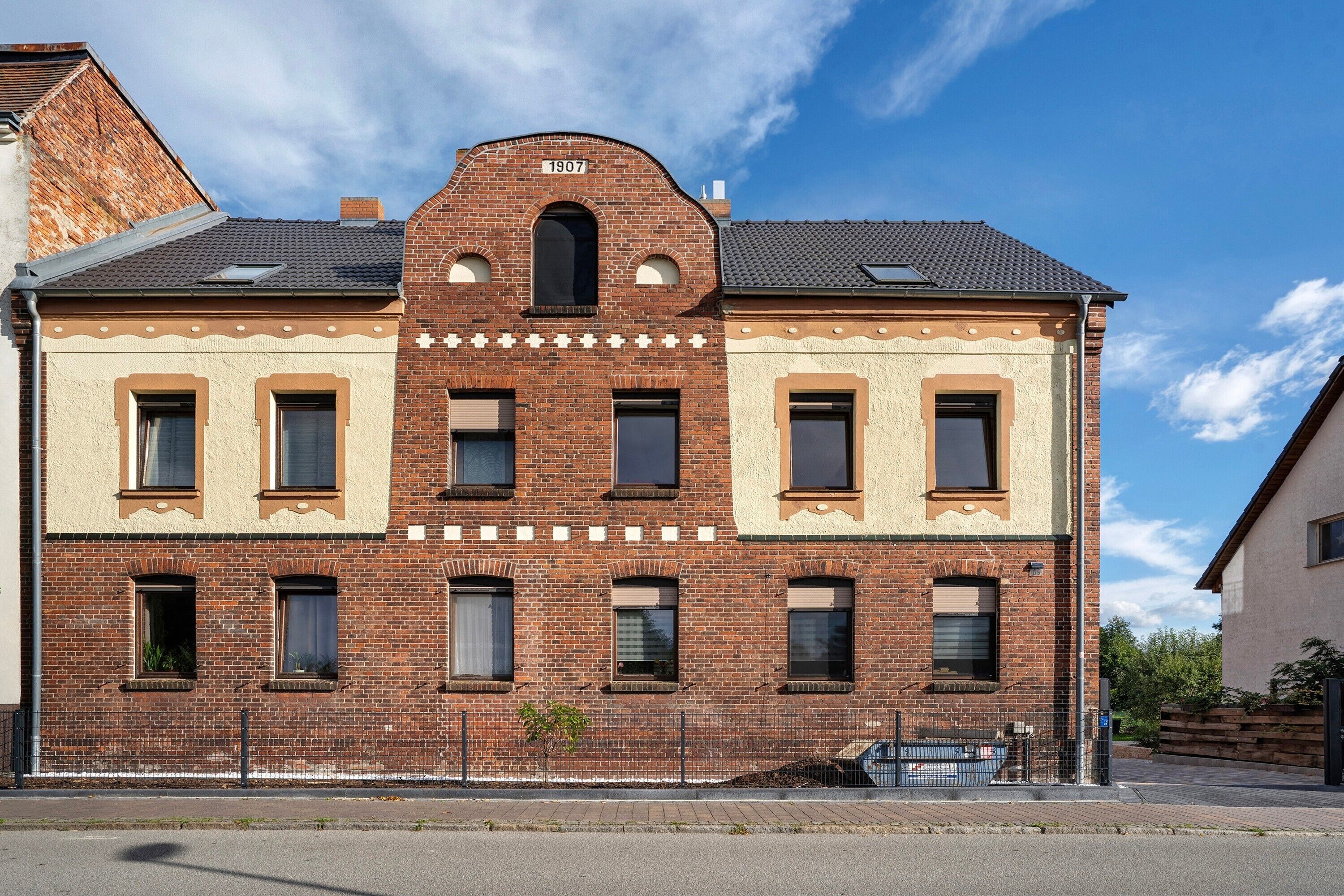 A two-story brick building with a flat roof, numbered 1907, featuring multiple windows and a small arched entrance.