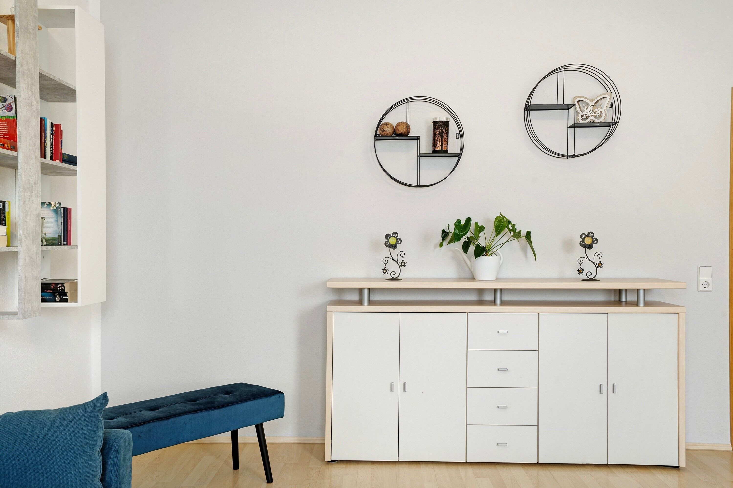 Una stanza con una libreria bianca, una panca azzurra, un mobiletto bianco con cassetti e due elementi decorativi circolari fissati alla parete.