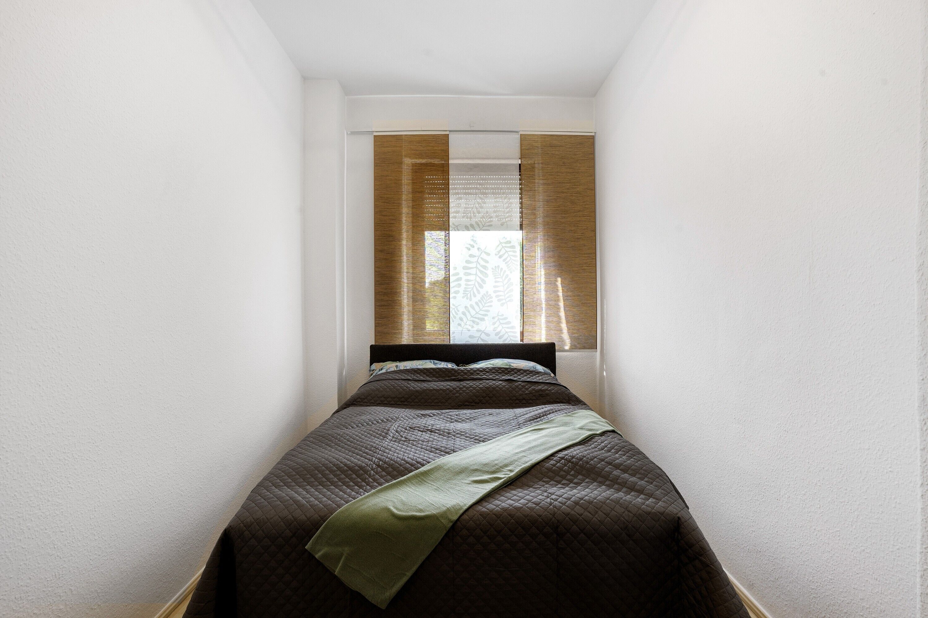 Ein schmaler Raum mit einem Bett, einem Fenster mit durchsichtigen Vorhängen und einer Tür.