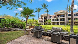 A patio area with two barbecue grills, a pond, and a building with balconies in the background. - Romantic Studio in Waikoloa Beach Resort (Waikoloa)