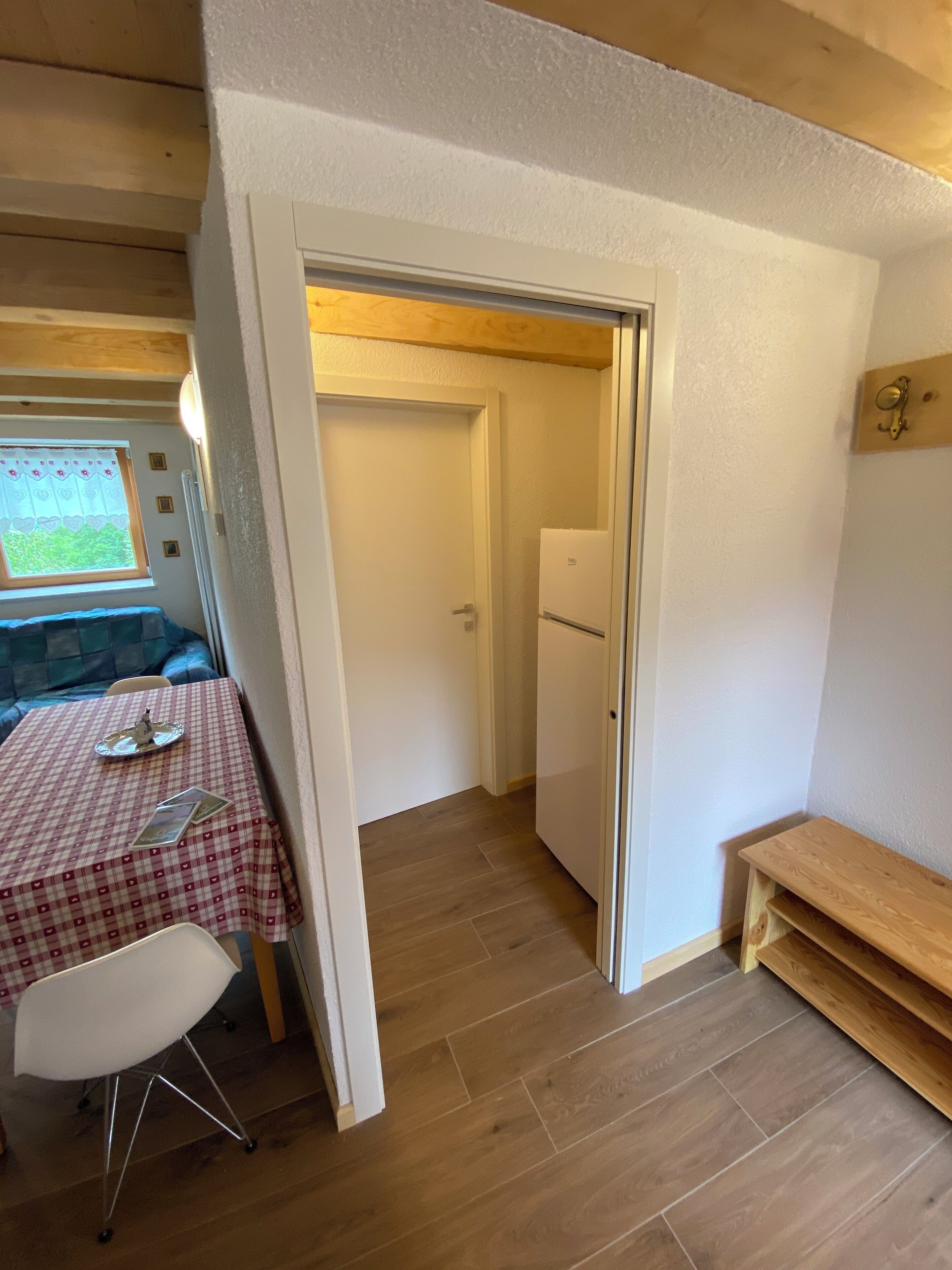 Ein kleines Zimmer mit Holzboden, einer weißen Tür, einem kleinen Tisch mit kariertem Tuch, einem weißen Stuhl und einer Holzbank.