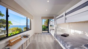 A modern bedroom with bunk beds, a window with a view of the ocean, and a small seating area with books. - Villa Luminosa in Mount Martha (Melbourne)