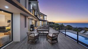 A modern house with a wooden deck, outdoor seating, and a view of the ocean at sunset. - Villa Luminosa in Mount Martha (Melbourne)