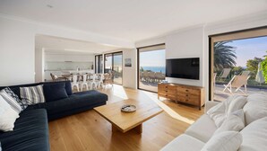A modern living room with a sofa, coffee table, and TV, large windows, and a balcony with outdoor furniture. - Villa Luminosa in Mount Martha (Melbourne)