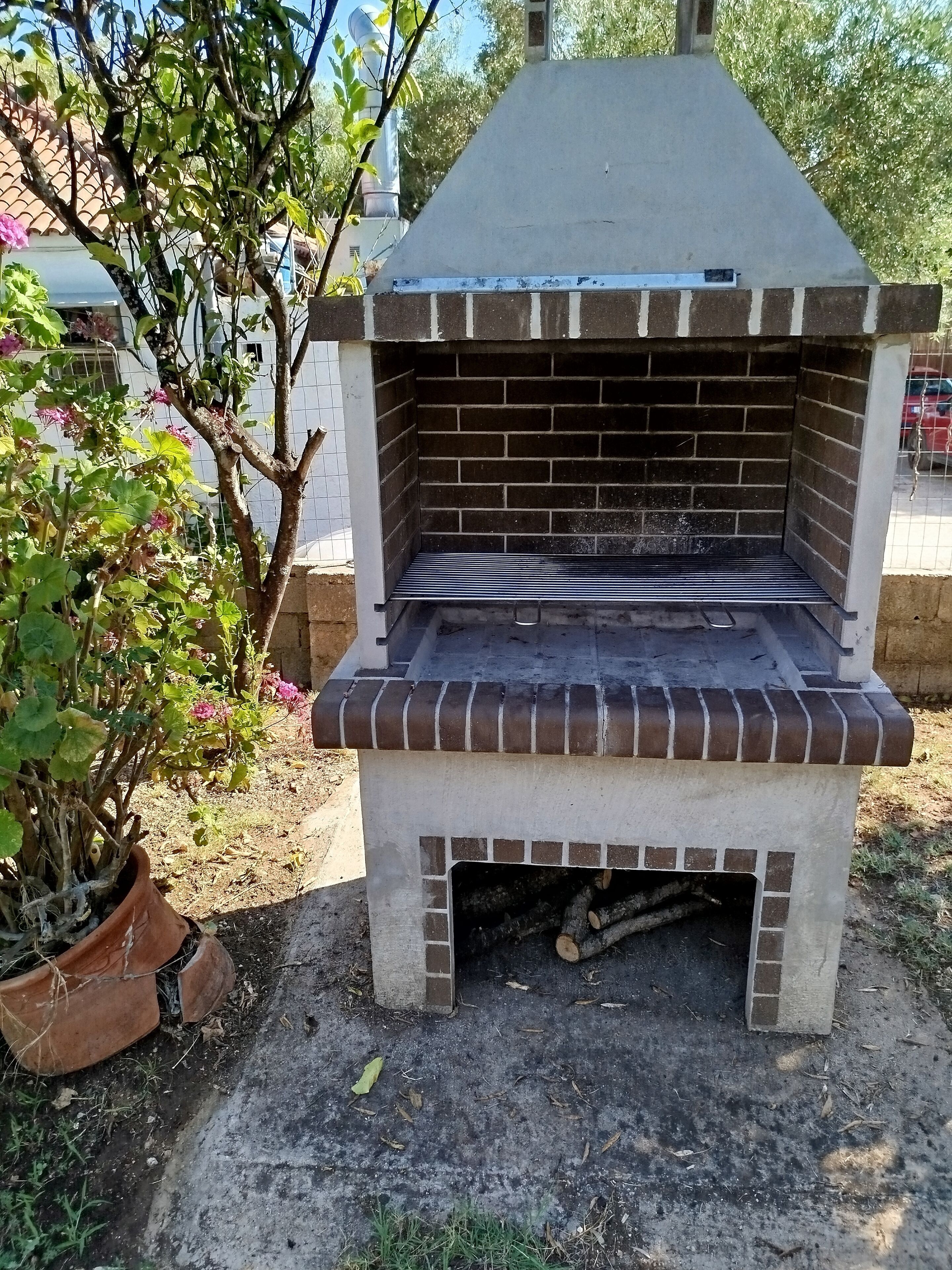 Un barbecue extérieur en briques, doté d’un four et d’une cheminée, entouré d’un jardin agrémenté de plantes en pot.