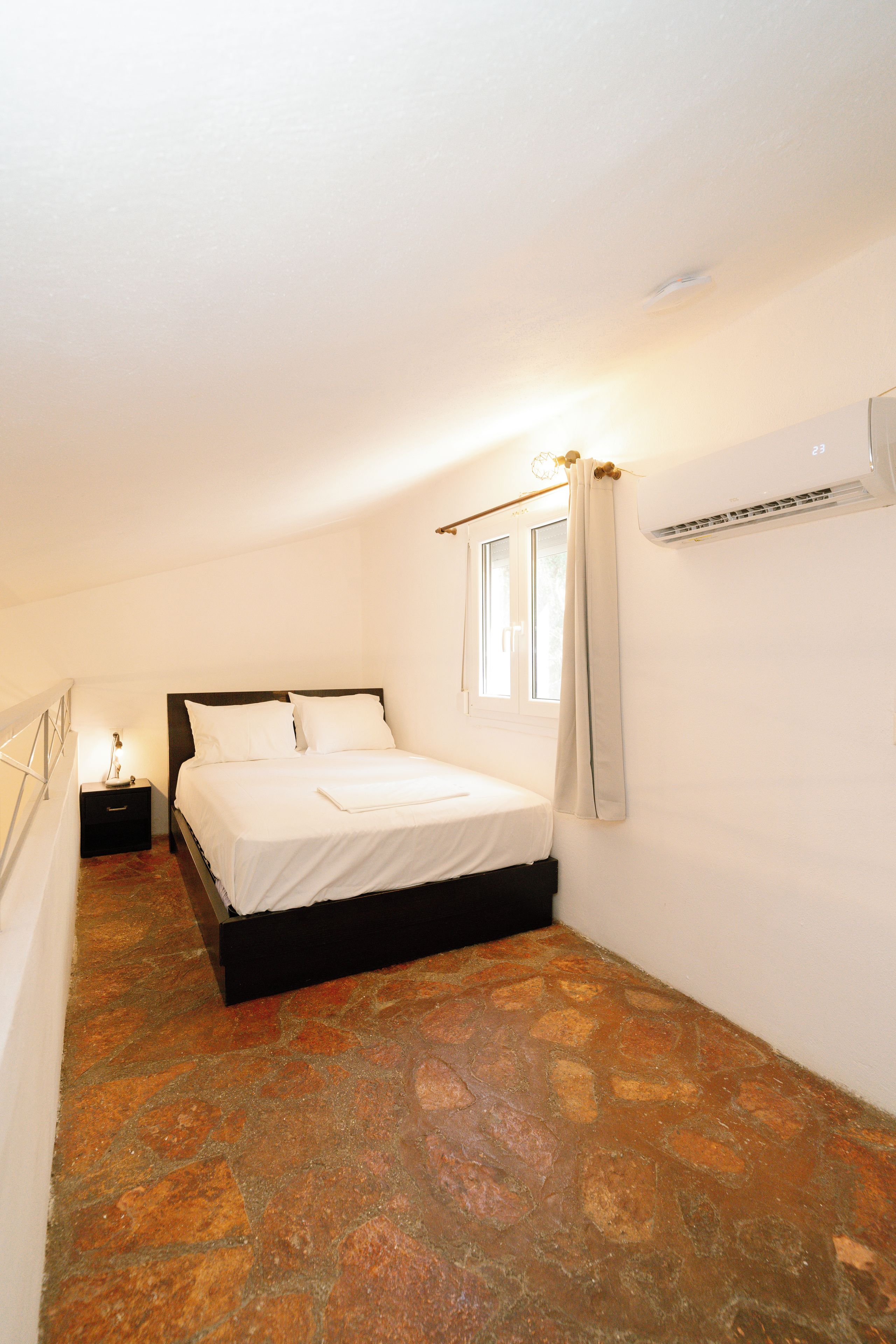Quarto com cama, mesa de cabeceira, janela com cortinas e ar condicionado.