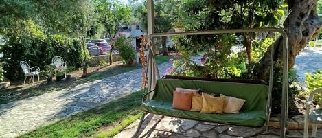 A covered outdoor seating area with a swing, stone flooring, and surrounding greenery.