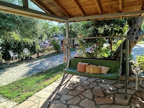 A covered outdoor seating area with a swing, stone flooring, and surrounding greenery. - Rozalia Studios  (Sivota)