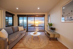 A modern living room with a sofa, a round coffee table, and a view of the sunset through large glass doors. - Azure Horizon Gas Heated Pool With Stunning View (Melbourne)