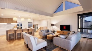 A spacious living area with a high ceiling, a kitchen with wooden cabinets, a dining area with chairs, a sofa set, and a television displaying the Netflix logo. - Azure Horizon Gas Heated Pool With Stunning View (Melbourne)