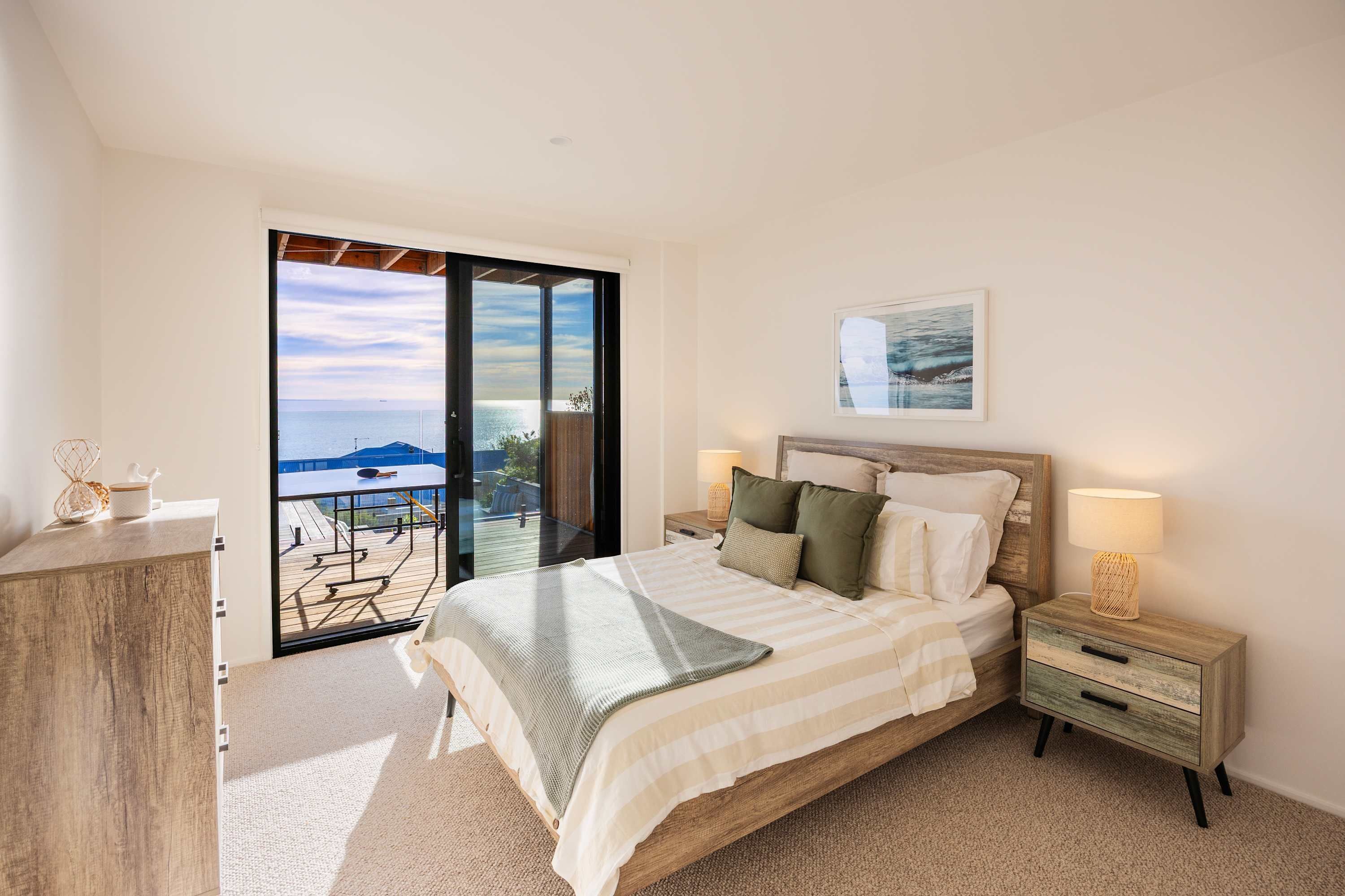 A bedroom with a bed, a nightstand, a dresser, and a view of the sea through a sliding glass door.