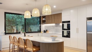 A modern kitchen with a central island, wooden chairs, and pendant lighting. - Azure Horizon Gas Heated Pool With Stunning View (Melbourne)