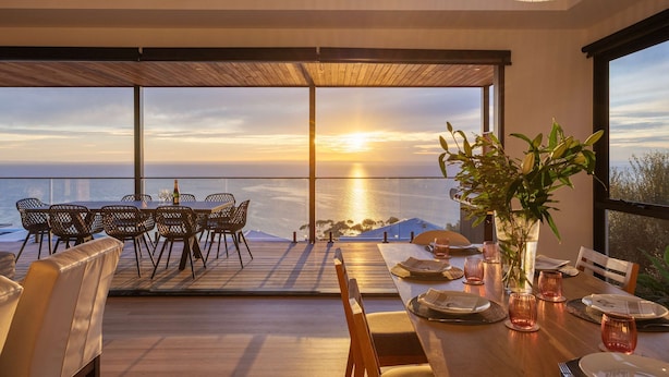 A dining area with a view of the sunset over the ocean, large windows, and a wooden deck. - Azure Horizon Gas Heated Pool With Stunning View (Melbourne)