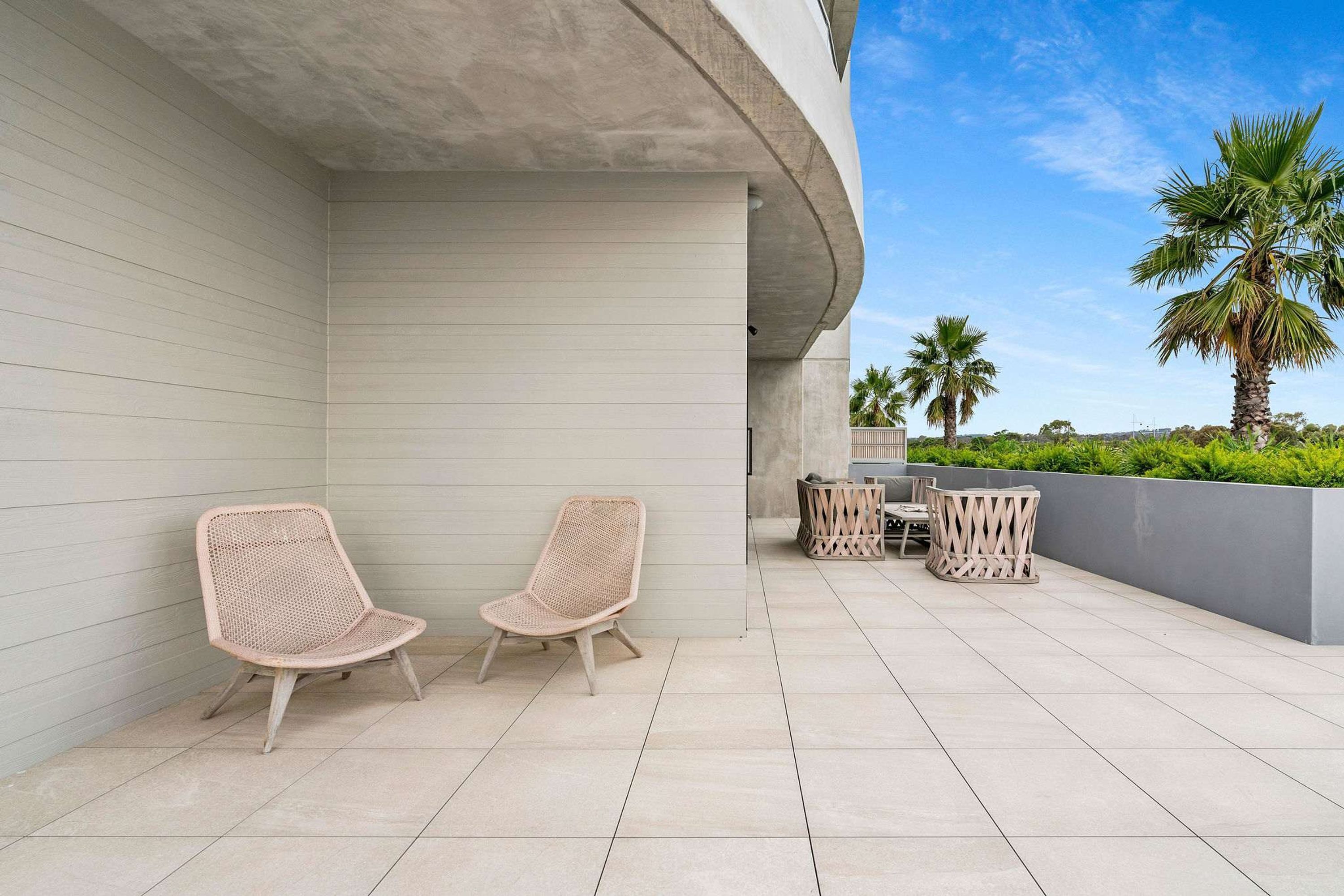 Une terrasse extérieure moderne, dotée de deux fauteuils en osier et d’une petite table, le tout entouré d’un muret et de palmiers.