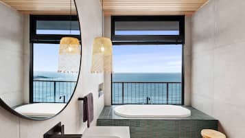 A modern bathroom with a large bathtub, a round mirror, and a view of the ocean through a large window.