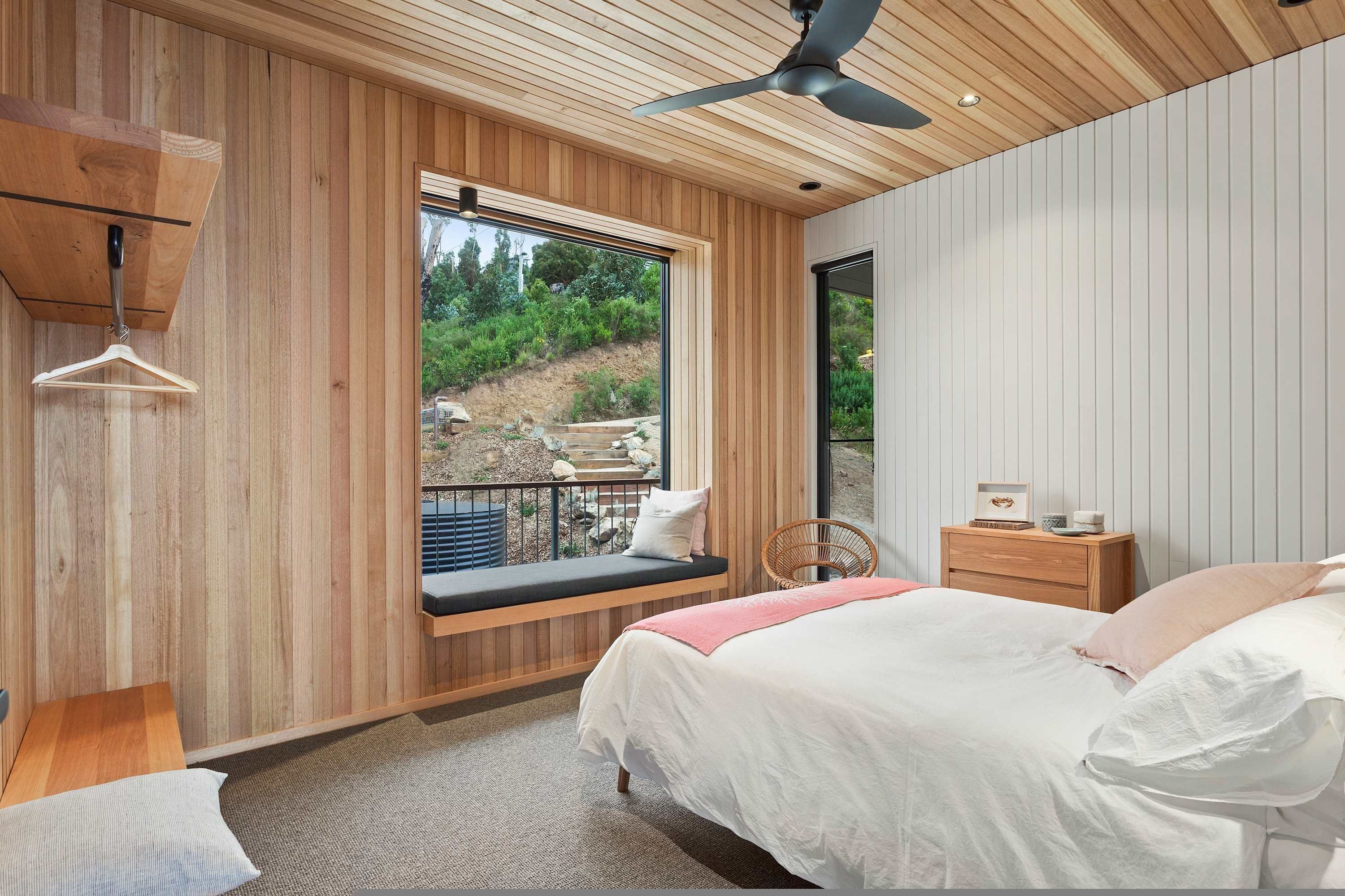 A bedroom with a large window, wooden walls, a bed, a bedside table, and a hanging light fixture.