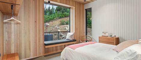 A bedroom with a large window, wooden walls, a bed, a bedside table, and a hanging light fixture.
