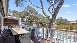 A deck with wicker furniture, a pool, and a view of trees and a building. - Saltwood Coastal Hamptons Luxe With Pool (Melbourne)