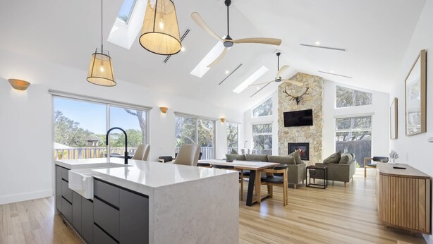 A spacious kitchen with a large island, a dining area with a table and chairs, a living room with a sofa and a fireplace, and a view of the outdoors through large windows. - Saltwood Coastal Hamptons Luxe With Pool (Melbourne)