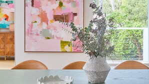 Un comedor con una mesa, sillas, un jarrón con plantas y un cuadro abstracto grande en la pared.