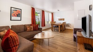 Ein modernes Wohnzimmer mit einer Couch, einem Couchtisch, einer Essecke mit Tisch und Stühlen sowie einem Fernseher auf einem Holzgestell.