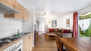Eine kompakte Küche mit Herd, Spüle und Holzschränken. Es gibt eine Essecke mit Tisch und Stühlen sowie ein Wohnbereich mit Sofa und Fenster.