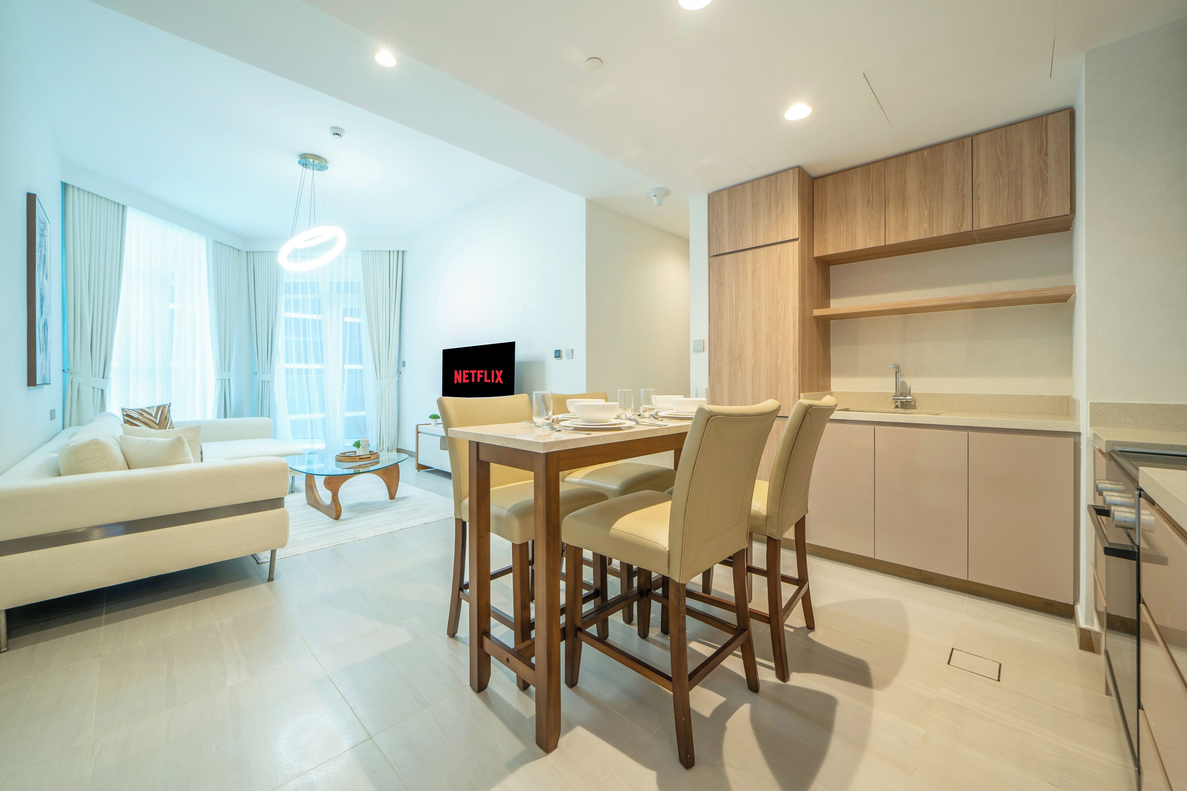 A modern living room with a sofa, dining table, and chairs, a kitchen area with wooden cabinets, and a TV displaying the Netflix logo.