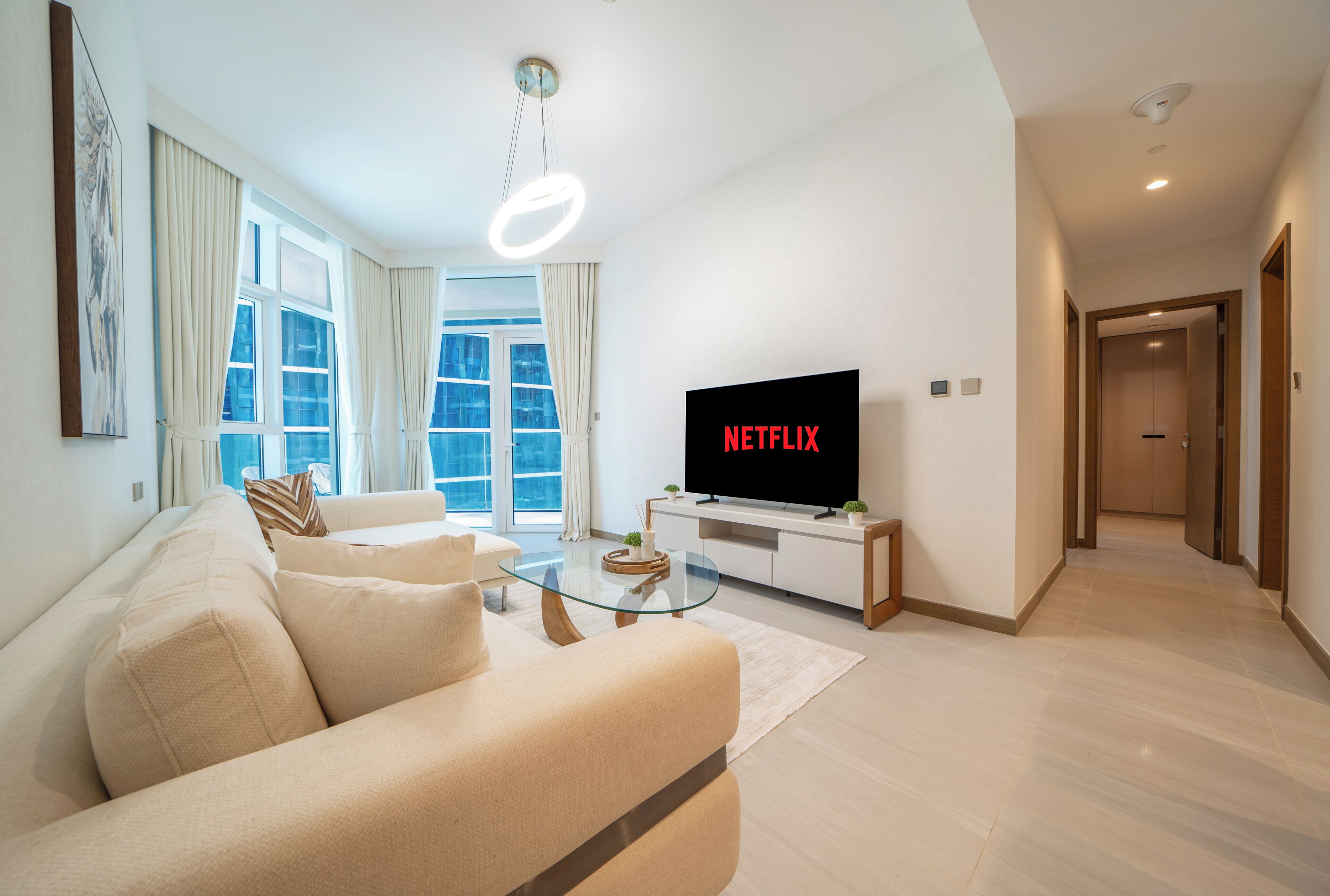A modern living room with a sofa, a glass coffee table, a flat-screen TV, and a hallway leading to a door.