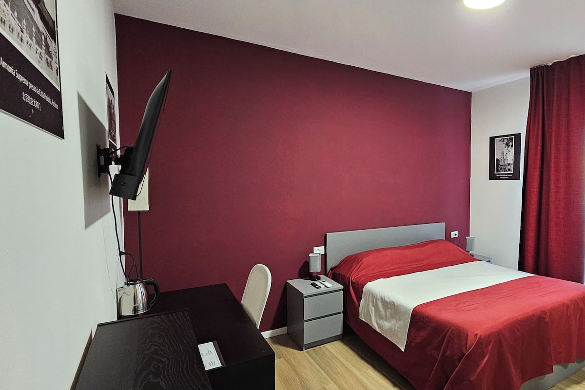 A bedroom with a bed, a desk, a chair, a nightstand, and a TV mounted on the wall.