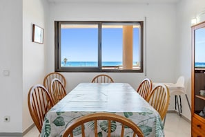 Un comedor con mesa y sillas, una ventana que ofrece vistas a la playa y un mueble con vajilla.