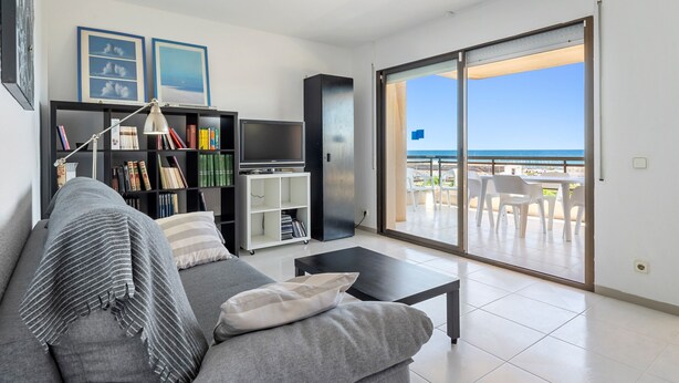 Un salon comprenant un canapé, une table basse, une bibliothèque, un téléviseur et une porte-fenêtre coulissante donnant sur un espace extérieur aménagé avec vue sur la mer.