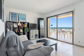 Una sala de estar con sofá, mesa de centro, estantería, televisor y puerta corrediza de vidrio que da a un área de estar exterior con vista al mar.