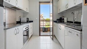 Une cuisine moderne avec des armoires blanches, un four encastré, un lave-vaisselle et une fenêtre donnant sur un balcon.