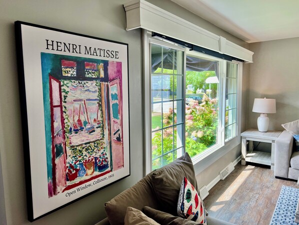 A living room with a large Henri Matisse painting, a window with a view of greenery, and a white lamp on a side table.