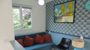 Una stanza con un divano blu, una sedia nera, un tavolino con una lampada, un quadro appeso al muro e una finestra vista alberi.