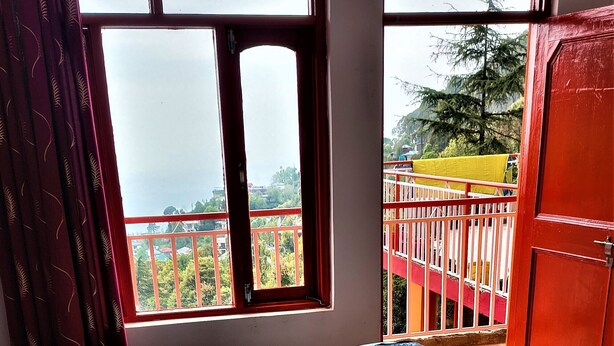 A bed with a blue sheet, a patterned rug, and a window with a view of a balcony and trees.