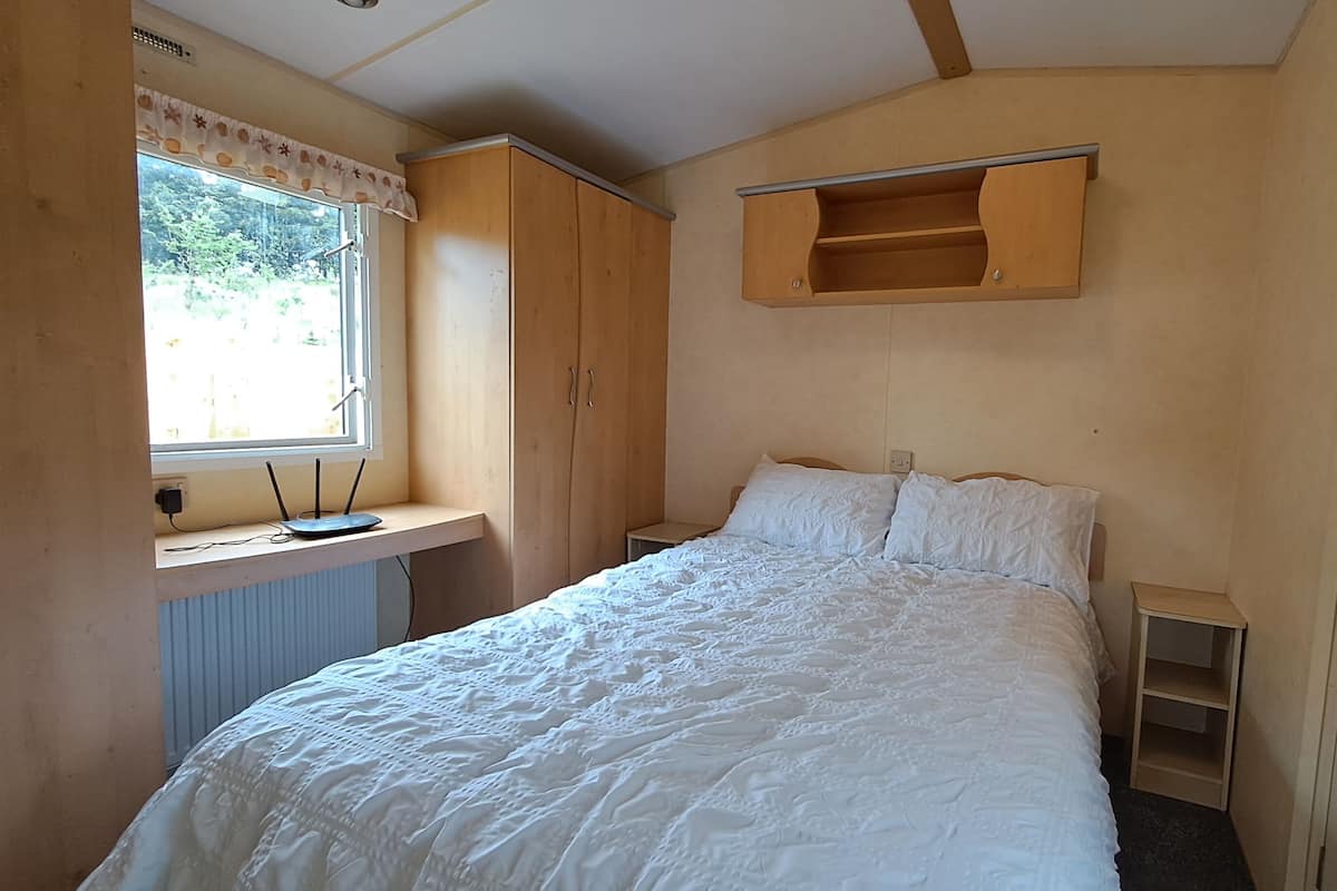 A bedroom with a bed, a window with a view, a wooden wardrobe, and a shelf above the bed.