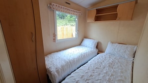 A small, cozy bedroom with two single beds, a window with a curtain, and a wooden shelf above the beds. - The Lookout - Park Home, Farm Park Stay (Swansea)