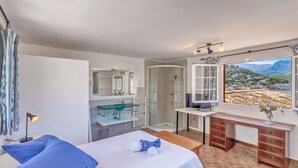 A bedroom with a bed, a desk, a shower, a bathroom with a glass sink, and a view of a mountainous landscape.