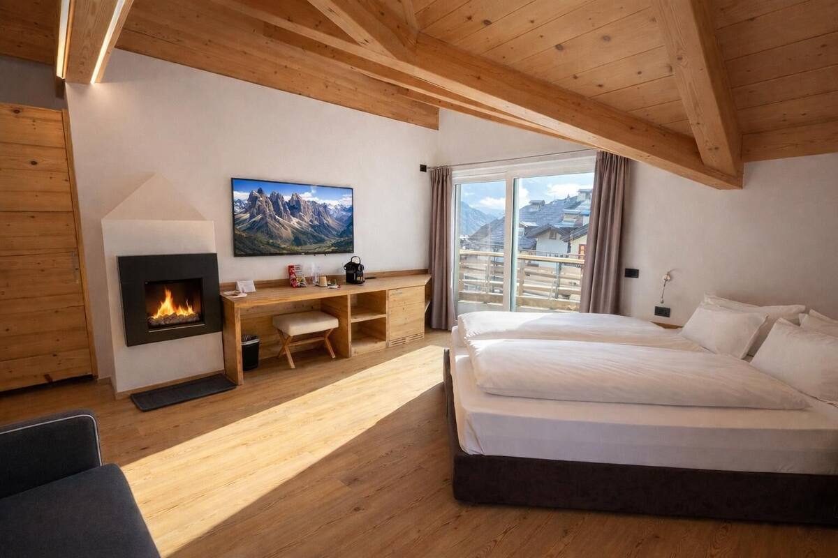 A bedroom with a bed, a desk with a chair, a fireplace, and a large window with a mountain view.