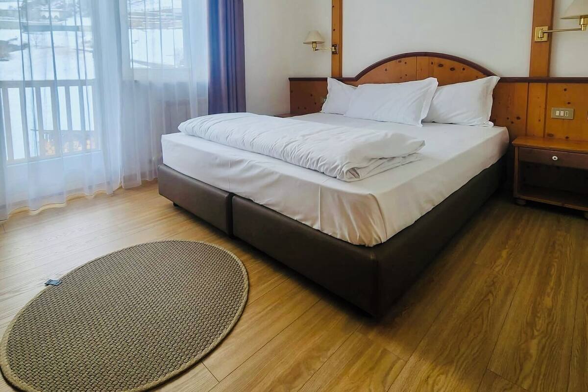 A hotel room with a bed, wooden headboard, nightstands, a round rug, and a window with curtains.
