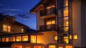 A modern building with a glass facade, wooden balconies, and two heated wooden cabins in the foreground. - HOTEL VILLA ROSA (Campitello di Fassa)