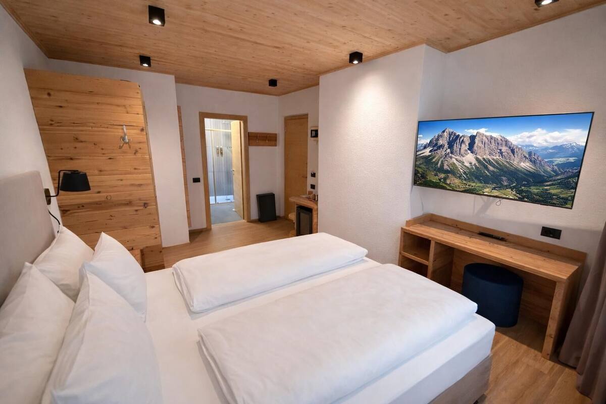 A hotel room with a wooden ceiling, a bed with white linens, a television on the wall, and a view of mountains.
