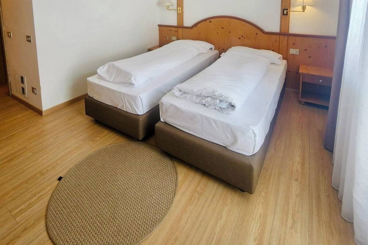 A hotel room with two single beds, a wooden headboard, a small bedside table with a lamp, and a round woven mat on the floor.