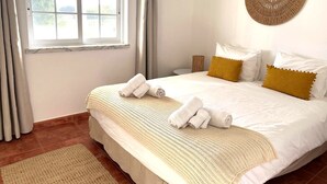Ein ordentlich bezogenes Bett mit weißen Leinen und zwei Kissen, ein gewebter Wandbehang und ein Fenster mit Vorhängen.