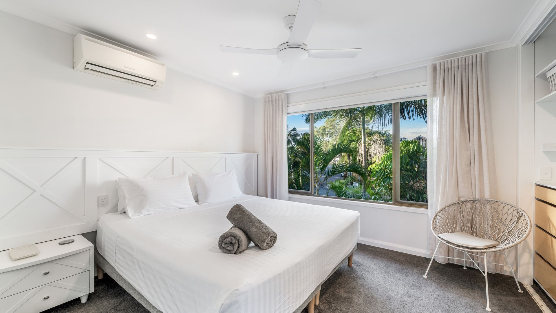 Camera d'albergo moderna con un letto grande, una ventola a soffitto, un condizionatore d'aria, una finestra con vista sul verde, un comodino bianco e una poltrona in vimini.