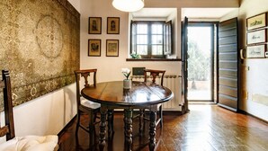 A dining area with a round wooden table, chairs, a hanging light fixture, and a window with open shutters.