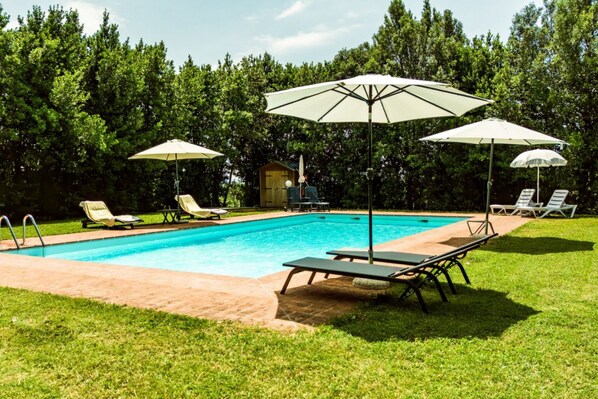 Piscina con sillas de descanso y sombrillas, rodeada de césped y árboles.