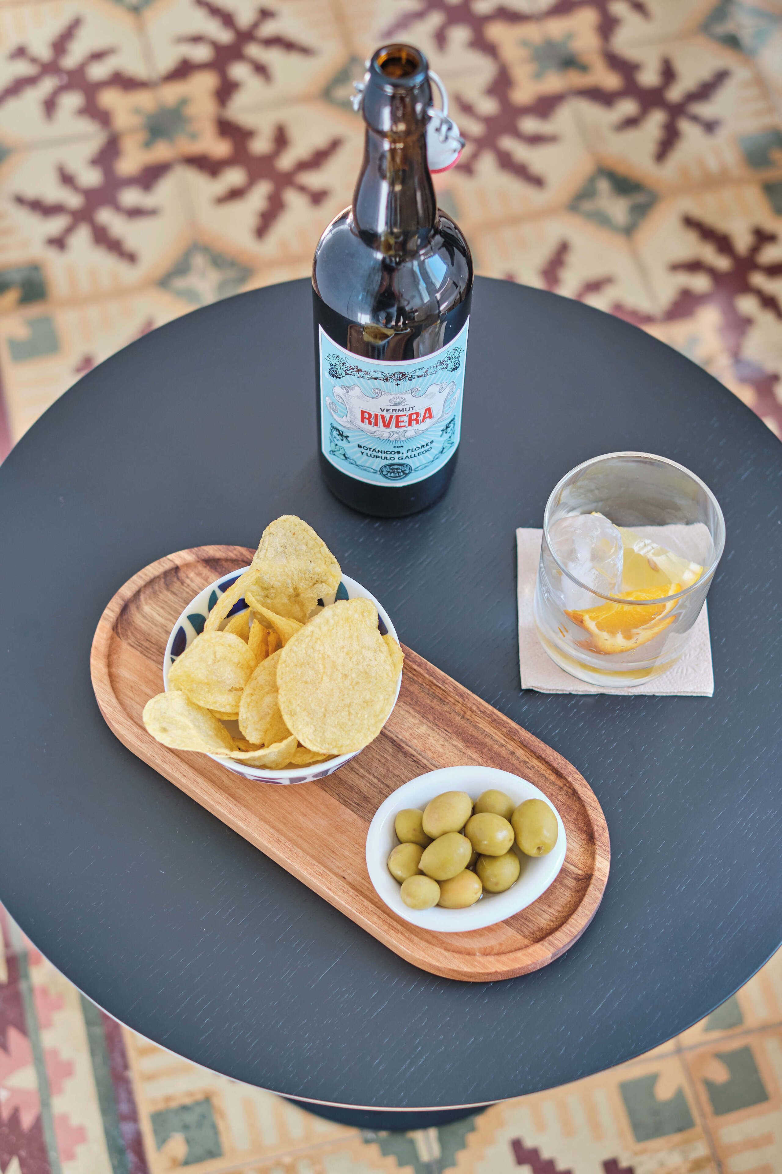 A bottle of Rivera wine, a bowl of chips, a glass with an orange slice, and a bowl of olives on a round table.