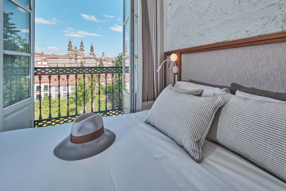Une chambre d’hôtel avec une grande fenêtre, un lit recouvert de draps blancs, un oreiller à rayures et un chapeau posé sur le lit.